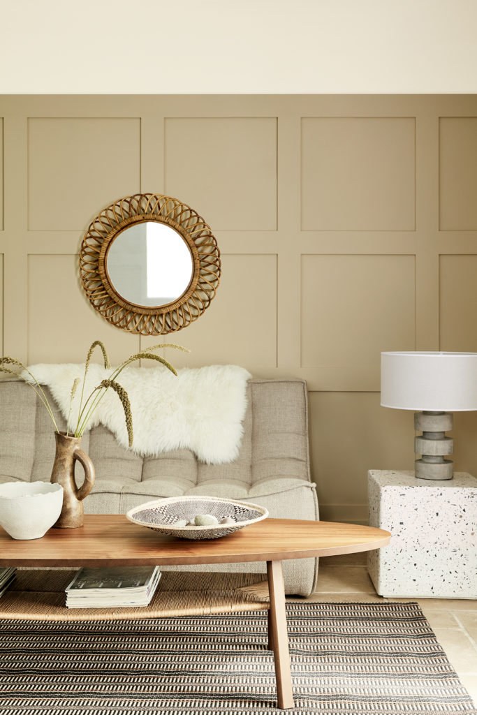 A cozy living room with beige paneled walls and warm neutrals. A round wicker-framed mirror hangs above a plush beige couch with a white fur throw. A wooden coffee table with decorative items sits on a striped rug. A modern lamp on a stone side table completes the minimalistic look.