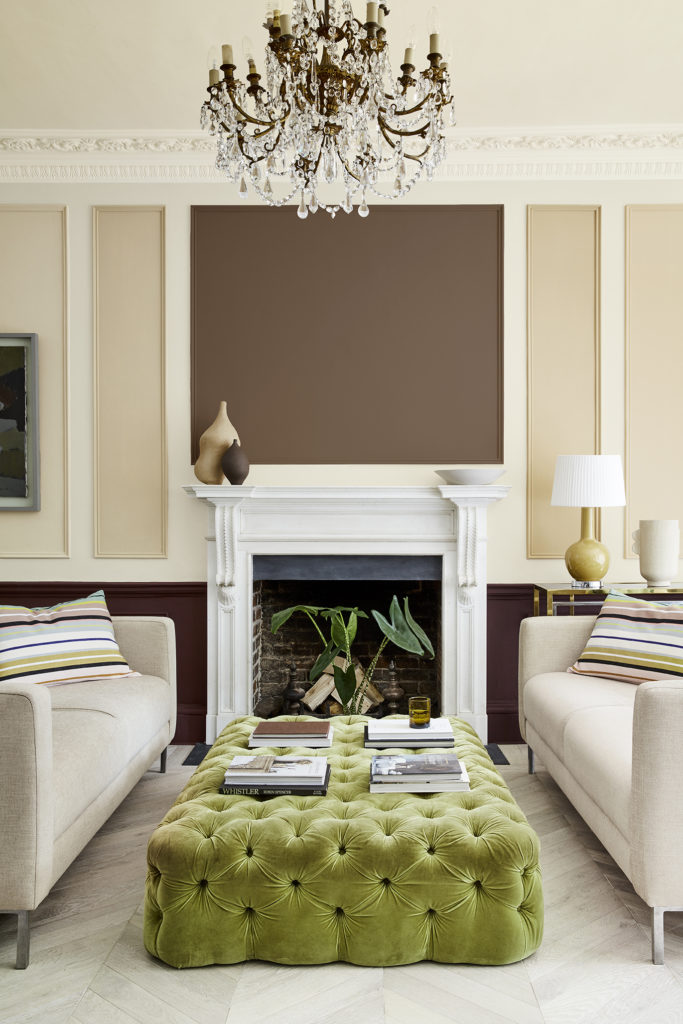 A sophisticated living room features an elaborate chandelier, a large tufted green ottoman with books and a drink placed on top, two beige sofas with striped cushions in warm neutrals, and a white marble fireplace. The mantel has art and a vase with a plant in front of the fireplace below.