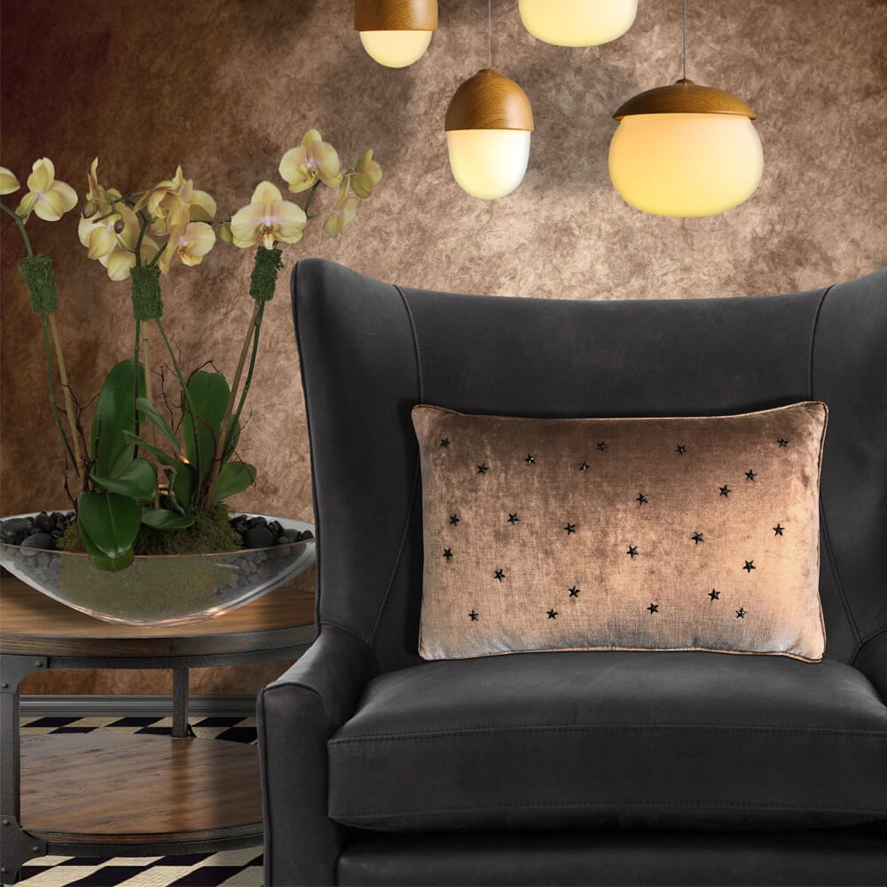 A cozy interior featuring a dark gray armchair with a beige star-patterned cushion. Above, three hanging pendant lights with warm glows. To the left, a modern table displays a shallow bowl with green ornamental plants and light yellow orchids on tall stems, all set against warm neutrals.