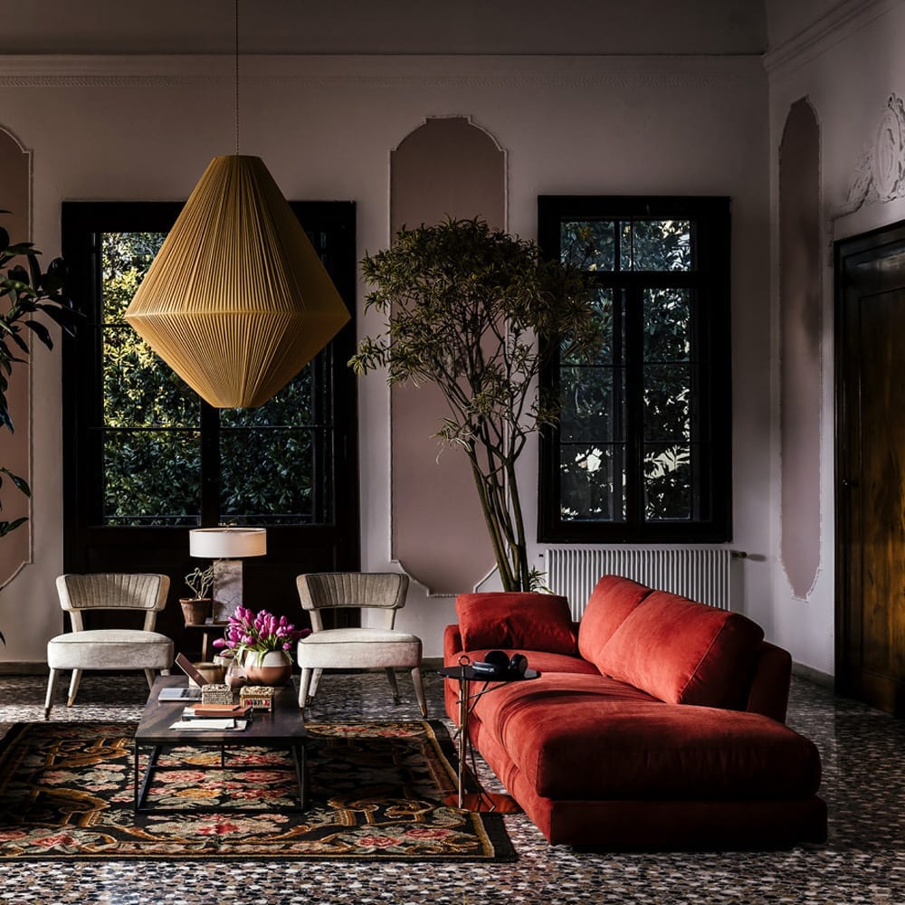 A stylish living room with a red velvet sectional sofa, two modern beige chairs in warm neutrals, a coffee table with decorative items, a large geometric chandelier, and floor-to-ceiling windows with dark frames. An indoor tree and vibrant patterned rug add to the cozy atmosphere.