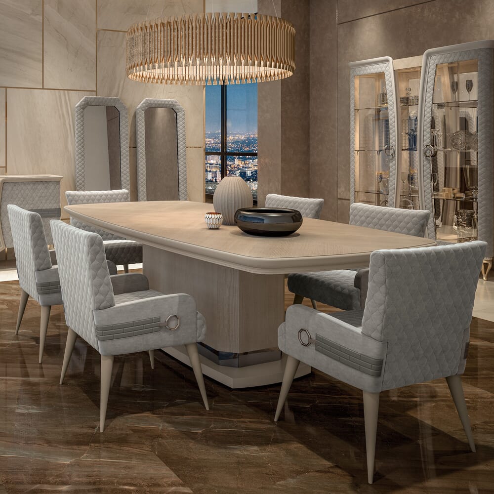 Modern dining room with a long, light wood table set with eight cushioned, grey quilted chairs. A wooden chandelier hangs above the table. The room features large windows, framed displays, and polished, glossy flooring in warm neutrals, with a cityscape view in the background.