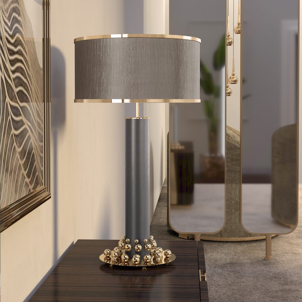 A modern table lamp with a gold and blue base, adorned with spherical accents, stands on a dark wood surface. The lamp, featuring a gray cylindrical shade with gold trim, complements the warm neutrals of the room. Behind it is an abstract art piece hanging on the wall and a tall mirror reflecting part of the space.