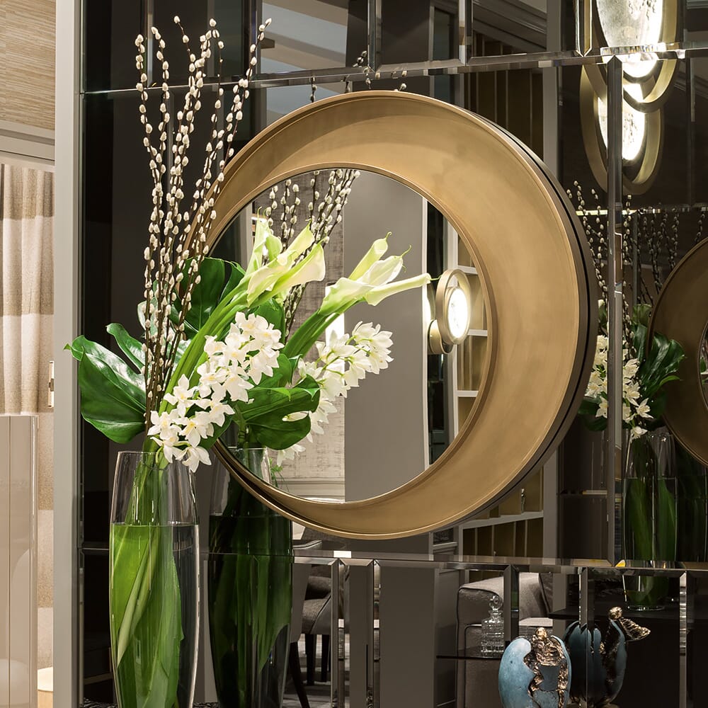 A stylish interior setup featuring a large decorative circular mirror with gold edging mounted on a reflective wall. In front of the mirror, there's a tall vase filled with white flowers and green leaves. The overall ambiance is elegant and modern, enhanced by carefully chosen statement furniture pieces.