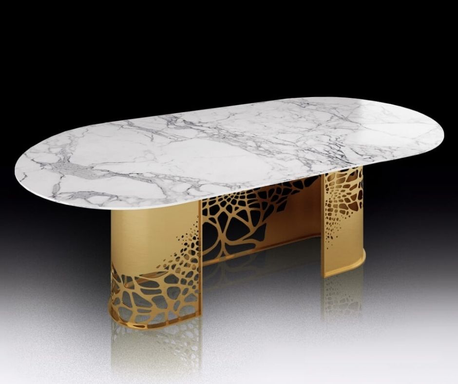 A modern, oval-shaped table featuring a polished, white marble top with gray veining. The table stands on two golden supports decorated with intricate, abstract cutout patterns, making it a stylish and elegant piece of statement furniture. The background is a simple gradient from black to gray.