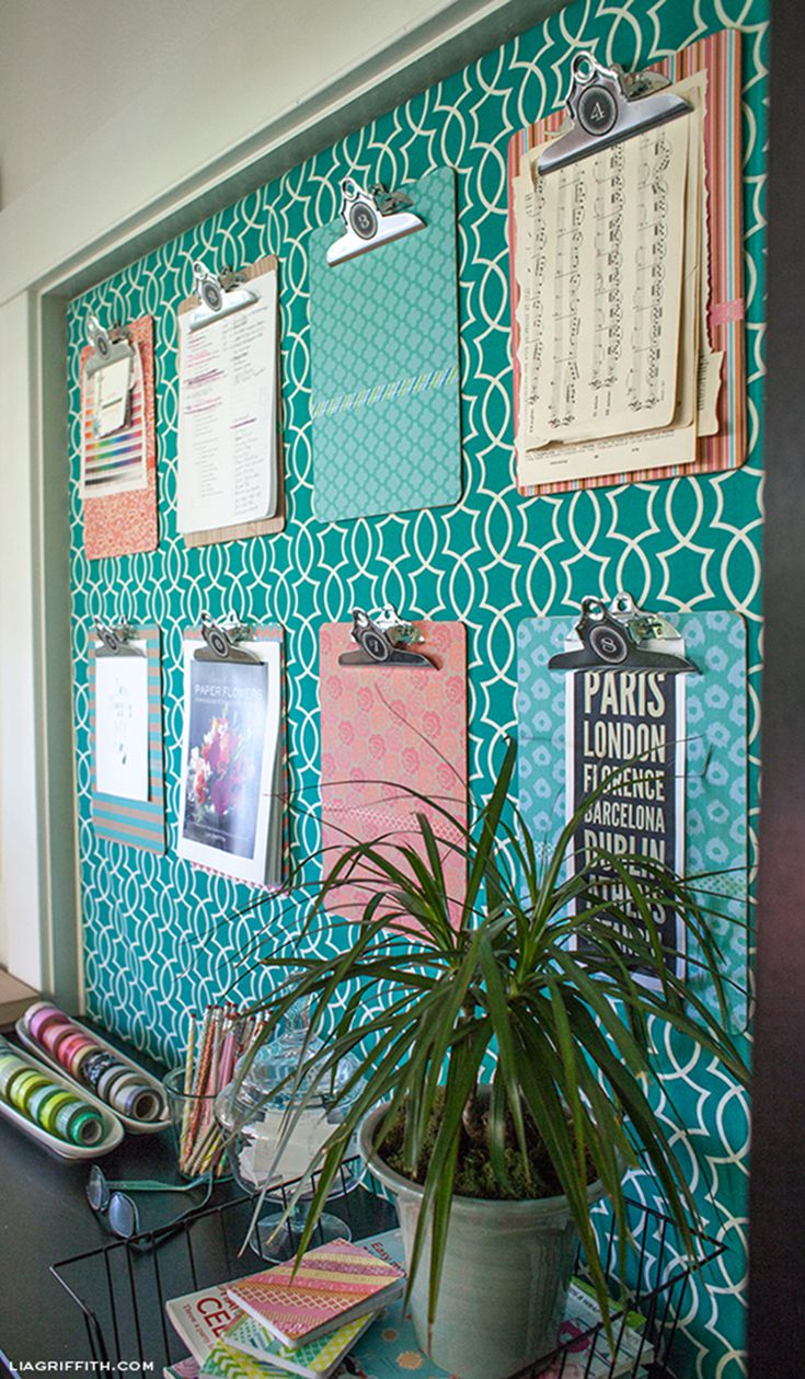 Style your home office with a wall decorated with six colorful clipboards holding various papers and art against a turquoise and white patterned background. Below the clipboards are rolls of patterned paper, books, and a green potted plant. The scene is bright and neatly arranged.