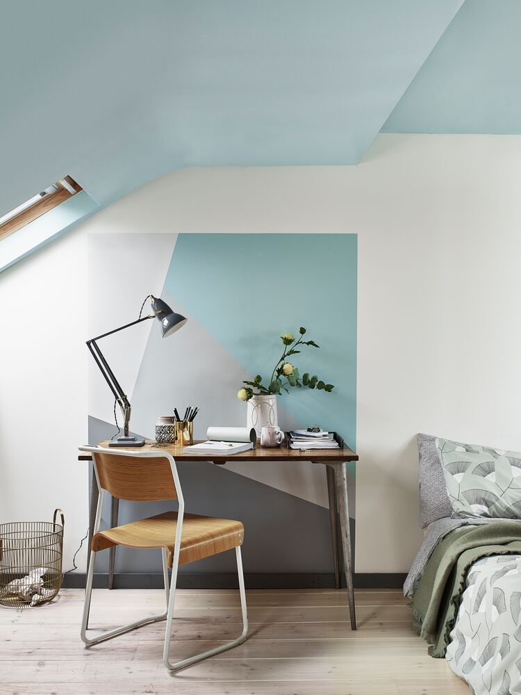 Style your home office with a modern workspace featuring a wooden desk and chair positioned under a slanted ceiling with a skylight. The desk holds a lamp, white vase with flowers, notebooks, and various stationery items. A bed with green linens is partially visible on the right.