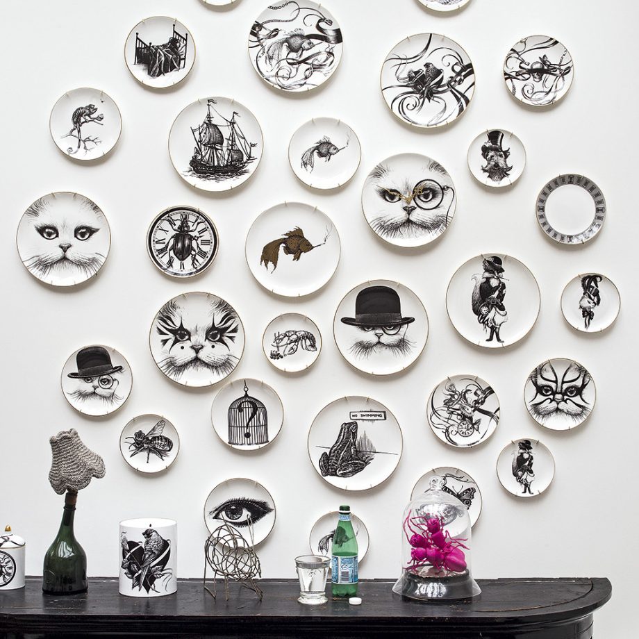 A white wall features an eclectic arrangement of decorative plates. Each plate displays a monochrome drawing, including eyes, cats, ships, insects, and Victorian-style figures. Style your home office with a black mantel below that holds various items, including a lamp, a bottle, and a pink flower.