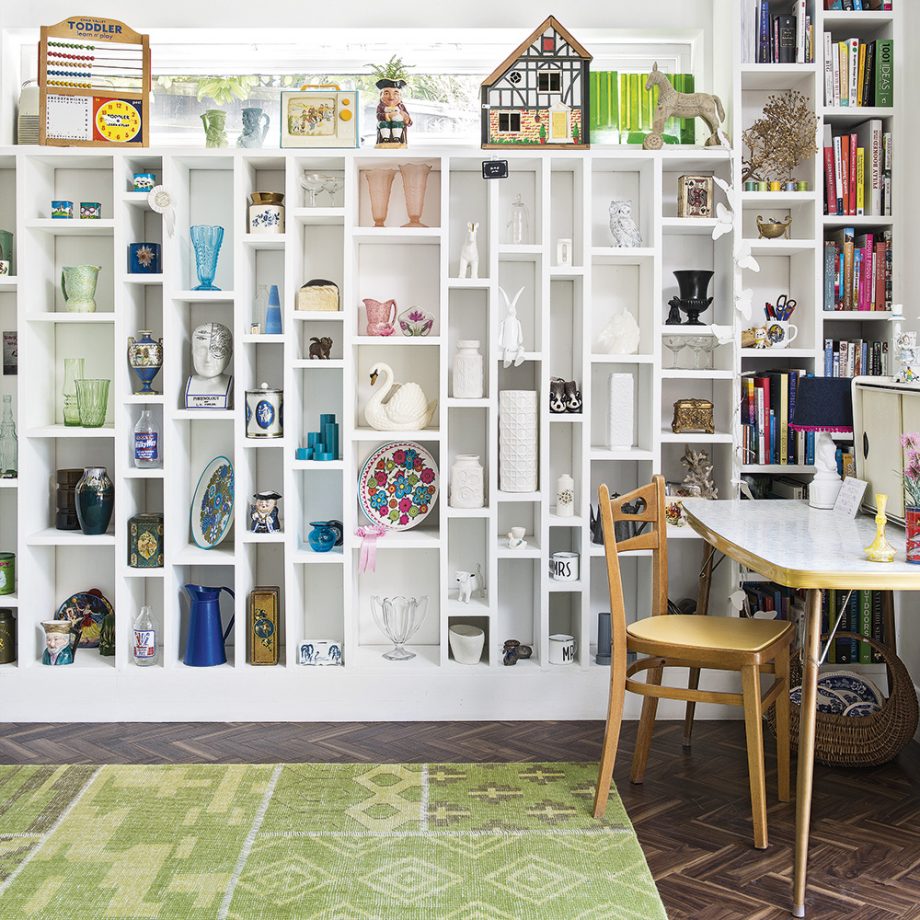 A bright room features a white wall with numerous shelves displaying various knick-knacks, figurines, glassware, and decor items—perfect to style your home office. A round table with a wooden chair sits to the right, while a green patterned rug covers the floor. Books fill several shelves to the far right.