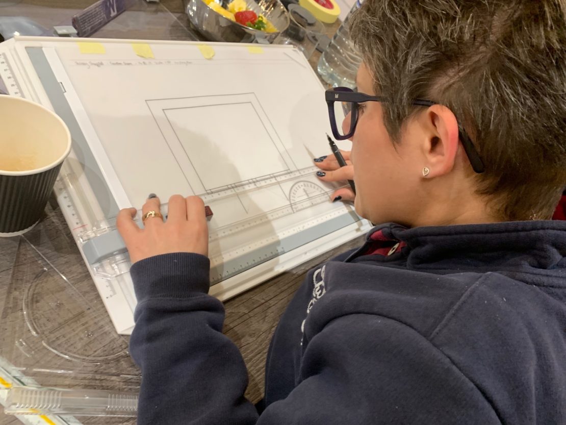 A person with short hair and glasses is focused on drawing a rectangular shape on a drawing board using technical tools, likely for an interior design course. A paper coffee cup is visible on the left, and colorful items are in the background. The person is wearing a dark sweatshirt.