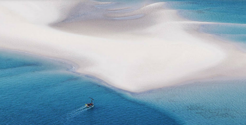 Aerial view of a small boat sailing in clear blue waters around a large, pristine sandbank. The stark contrast between the deep blue sea and the white sand creates a stunning, serene landscape, ideal for hosting exclusive luxury events.