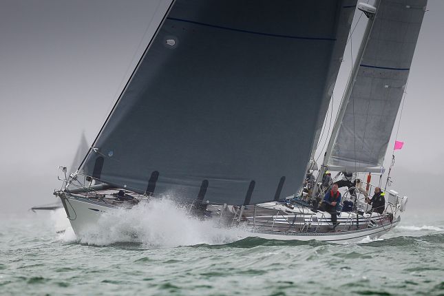 A sailboat with a dark sail cuts through choppy waters, creating a spray at the bow. The scene appears overcast with minimal visibility due to a misty atmosphere. Despite the conditions, this vessel is often utilized for luxury events, with the crew dressed in wet weather gear.