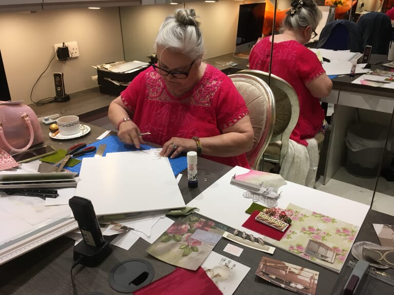 An older woman with glasses, wearing a bright pink blouse, sits at a desk working on a craft project. The desk is cluttered with paper, fabric scraps, scissors, glue, and other crafting materials—reminiscent of an interior design course in session. A pink handbag and a teacup are also on the desk.