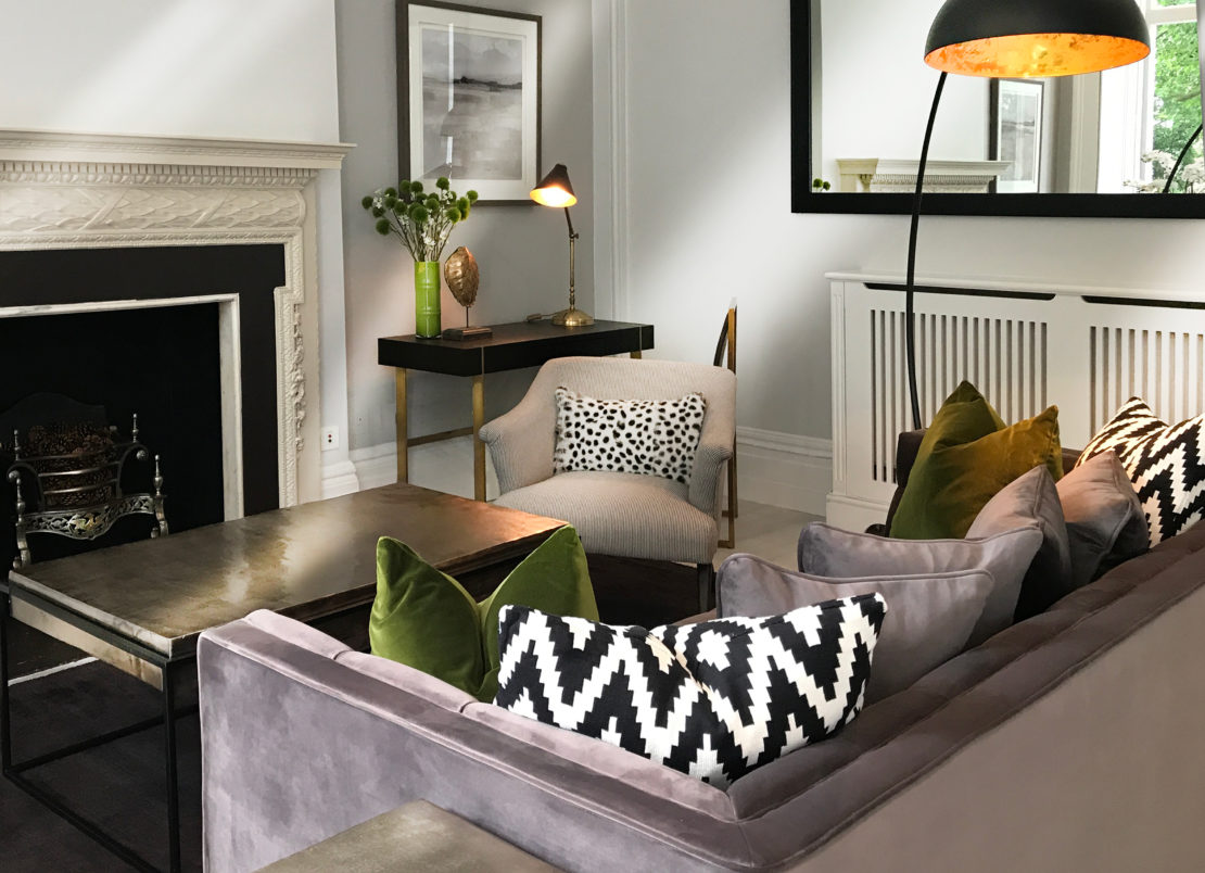 A modern living room with gray walls, a fireplace, and a large mirror showcases what you could learn in an interior design course. A gray couch decorated with green and geometric black-and-white pillows faces a metallic coffee table. A reading chair, a desk with a green vase, and a black floor lamp complete the decor.