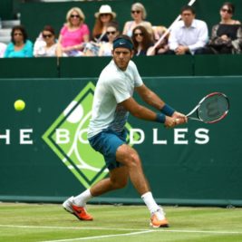 A tennis player in action on a grass court, preparing to hit a backhand shot. He is wearing a white shirt, teal shorts, and orange sneakers. Spectators sit in the background, watching the match at one of the season's luxury events, with a green banner behind the player displaying "The Boodles" logo.