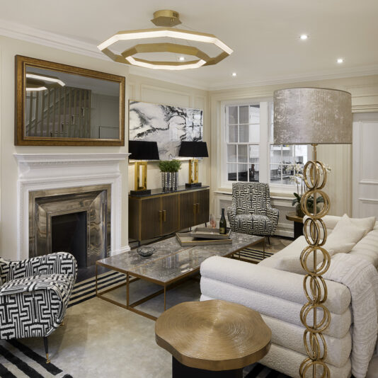 A modern, stylish living room featuring a geometric-patterned armchair, a plush white sofa, a marble coffee table, and a fireplace with a large mirror above. A unique lamp with circular accents stands next to the sofa. A staircase with beige carpet is seen on the right.