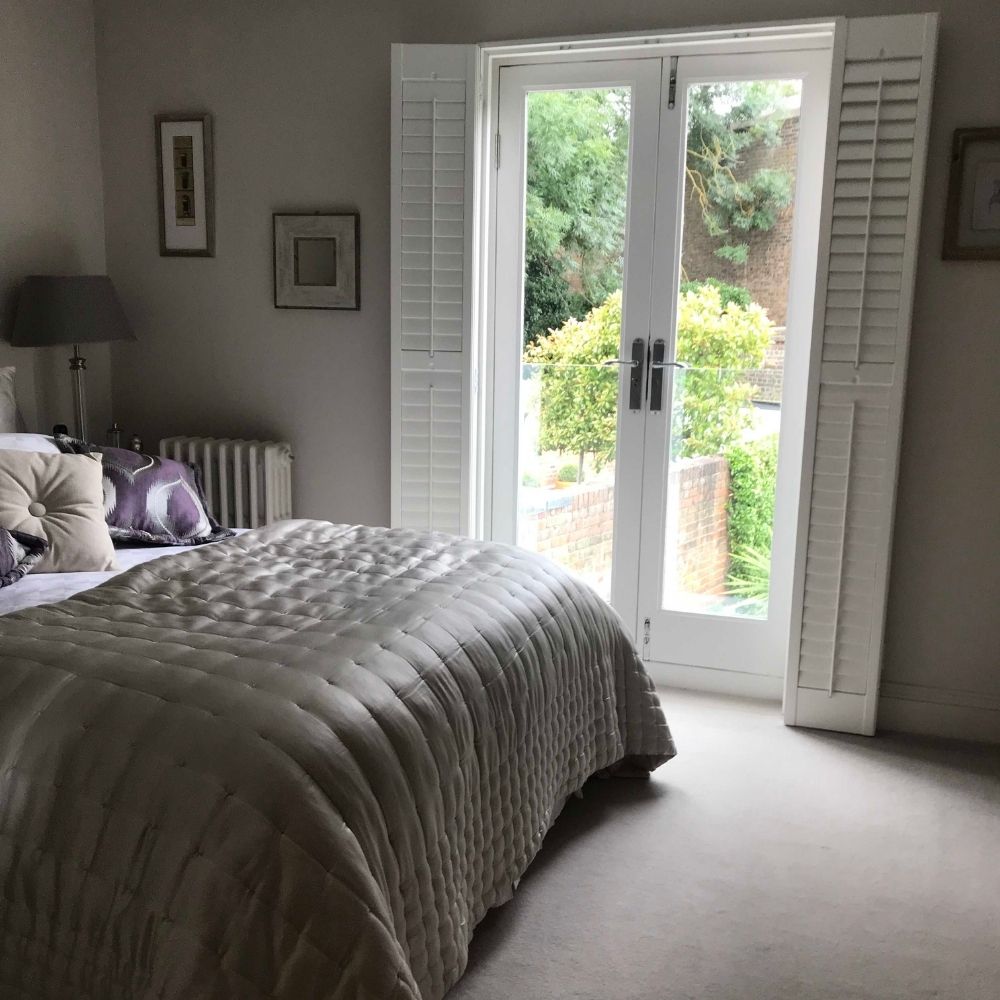 A cozy bedroom featuring a neatly made bed with a light-colored quilt, a side table with a lamp, and framed artwork on the walls. French doors with white shutters open to a view of a green garden area outside. This well-planned interior design project includes carpeted flooring that adds to the room's warmth.