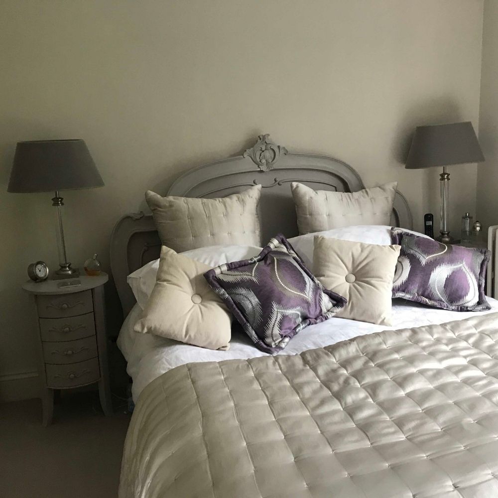 A neatly made bed with a tufted beige headboard, beige and purple patterned pillows, a beige quilt, and bedside tables with matching lamps on either side completes this elegant interior design project.