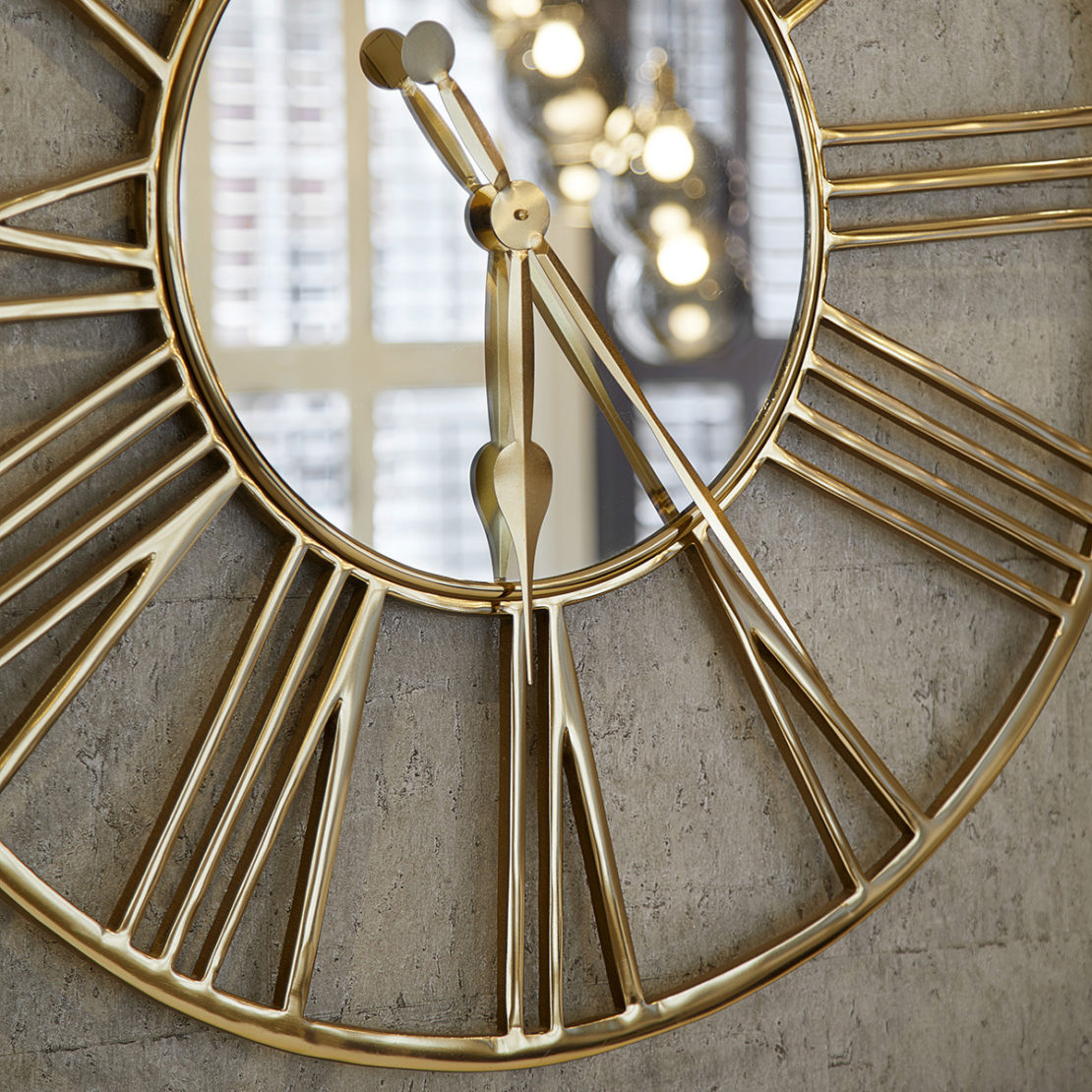 A close-up of a large, round wall clock with an intricate gold frame. The clock features Roman numerals and gold clock hands. The reflective surface captures blurred lights in the background, adding a warm and elegant ambiance—perfect for any interior design project.