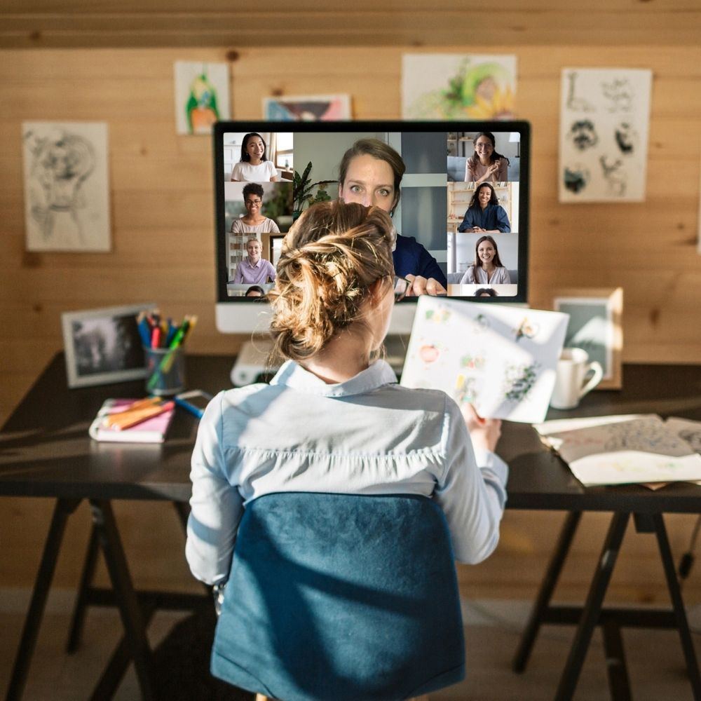 A person with their back to the camera is sitting at a desk, participating in an online interior design course on a computer. The monitor displays several people in a virtual meeting. The desk is cluttered with papers, books, and stationery, in a room with wooden walls.