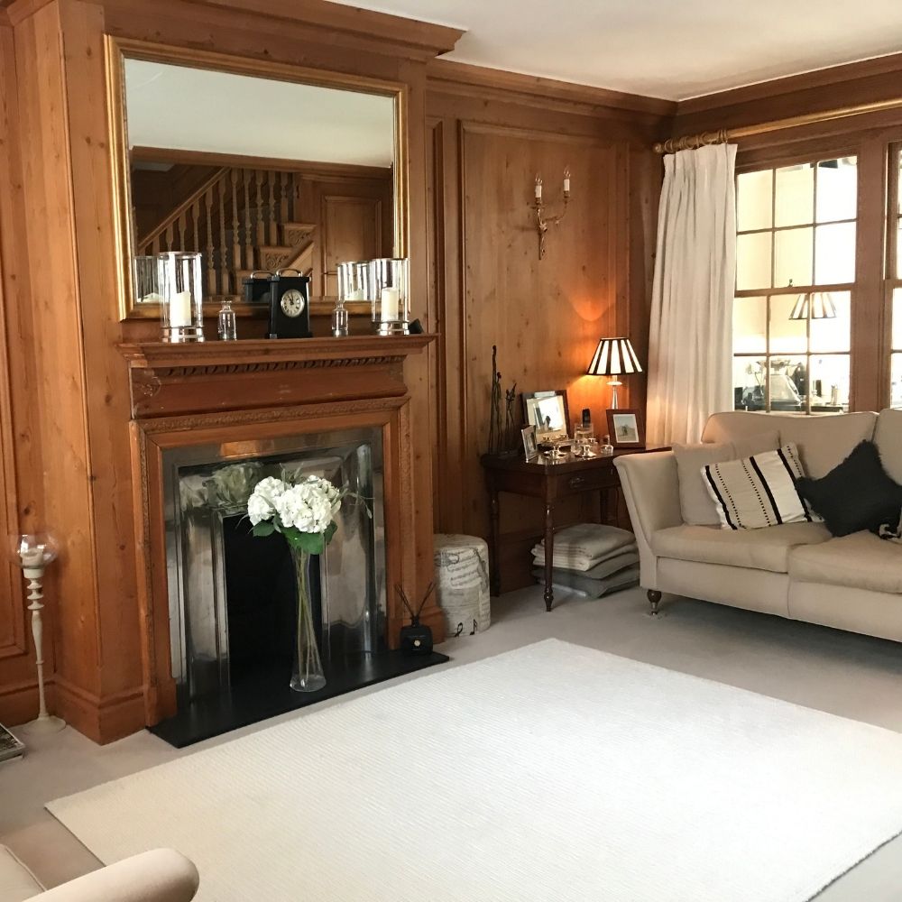 A cozy living room, part of an interior design project, features wood-paneled walls, a large mirror mounted above a fireplace, and a vase with white flowers on the hearth. A cream-colored rug covers the floor, and the furnishings include cream sofas with striped pillows and a side table with a lamp.