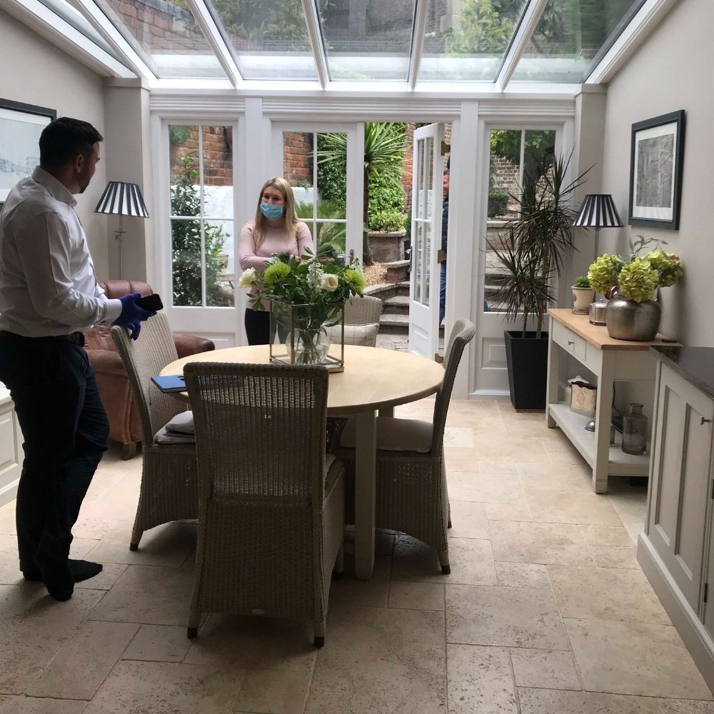 A man and a woman, both wearing face masks, stand in a bright room with a glass ceiling and large windows. The space, evidently part of an interior design project, is furnished with a round table surrounded by wicker chairs, a sideboard, and various plants.