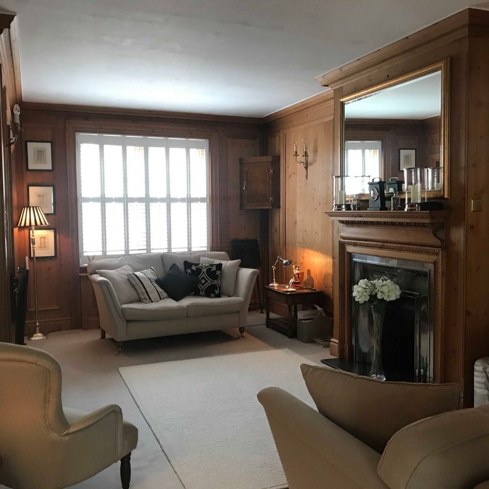 A cozy living room with wood-paneled walls, featuring a sofa with decorative pillows, an armchair, and a cushioned chair. A large mirror is above a fireplace adorned with candles, and a window with shutters lets in natural light. This inviting space is part of an exquisite interior design project.