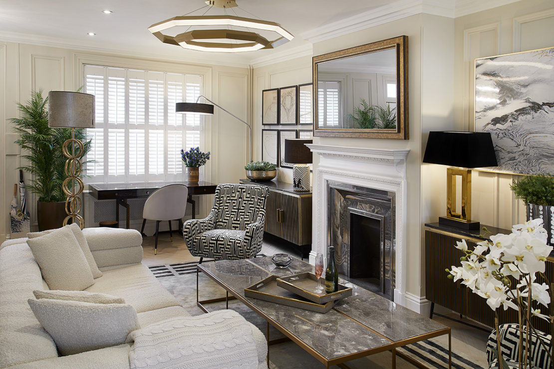 An elegant living room showcases a white sofa, patterned armchairs, and a marble coffee table. At the heart of this interior design project is a fireplace adorned with a large mirror, flanked by modern lamps and decorative items. A desk near the window and various artworks complete the aesthetic.