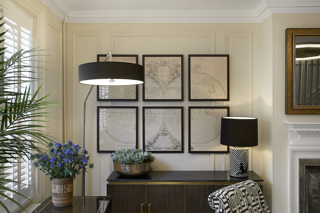 A stylishly decorated room, part of an impressive interior design project, showcases a wall featuring six framed pieces of art arranged in a grid. A tall floor lamp and a table lamp with black shades provide lighting. Below, a cabinet holds a potted plant and a small vase with purple flowers. A mirror graces the right wall.