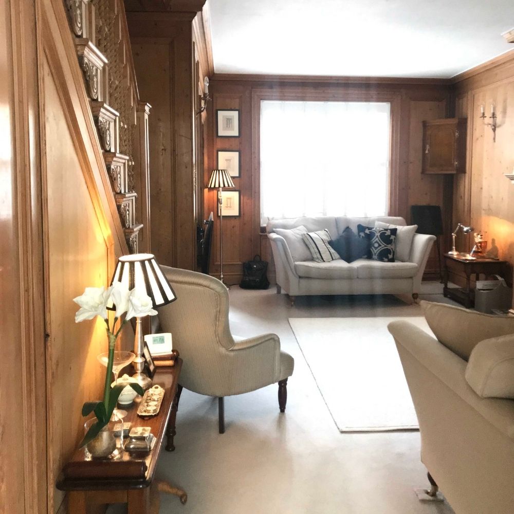 A cozy living room with wooden paneled walls, beige armchairs, and a sofa adorned with pillows. A side table with a lamp and vase of flowers is on the left, basking in light streaming through a large window. This inviting interior design project also features a staircase to the left.