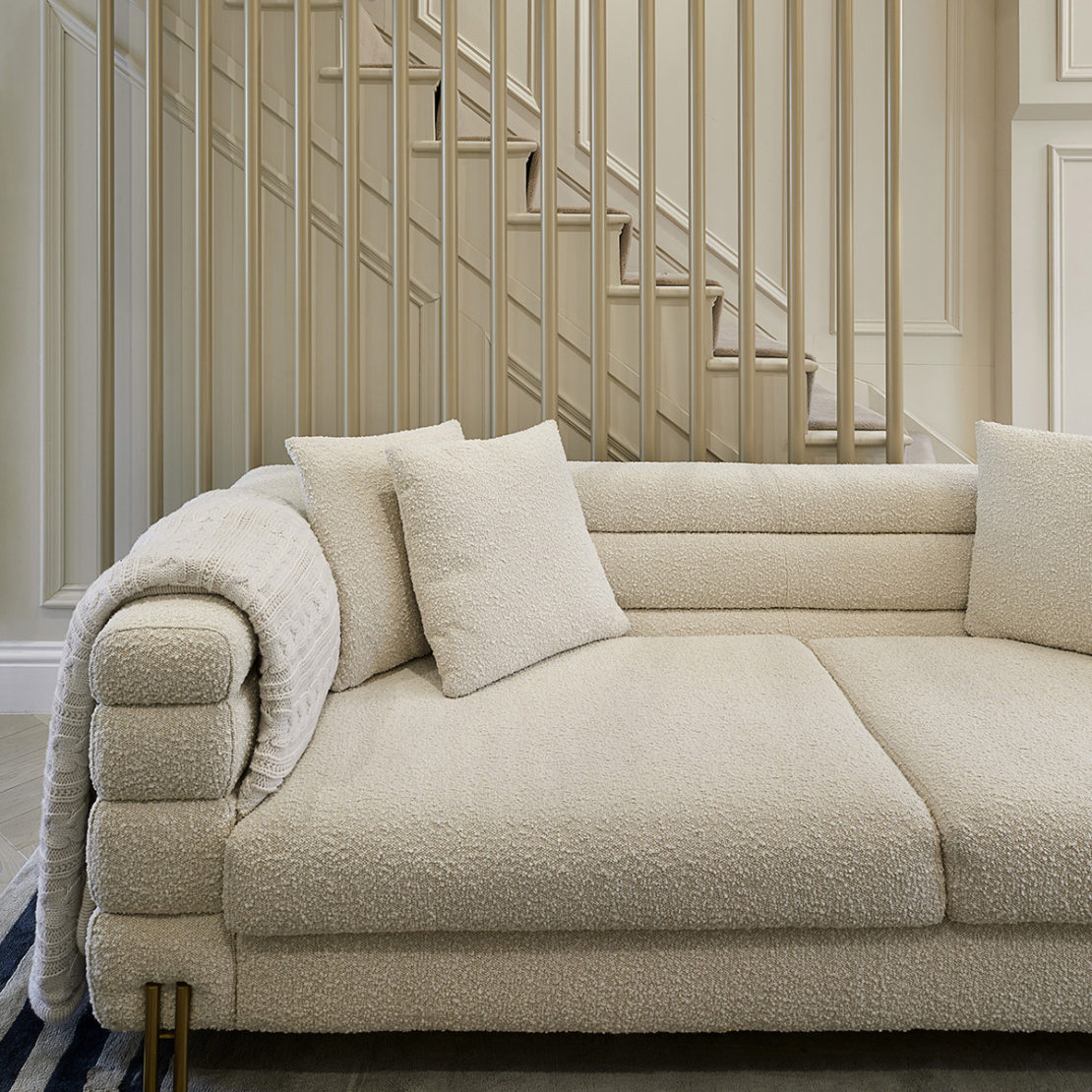 A modern, beige sofa with plush cushions and a rolled armrest sits in front of a staircase with vertical wooden railings. This interior design project boasts a simple yet elegant design, complementing the neutral tones of the room to create a cozy and inviting atmosphere.