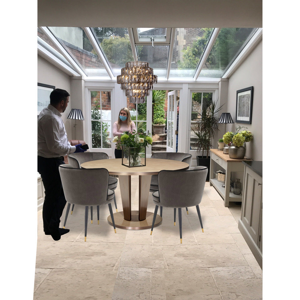 A modern dining room with a glass ceiling and light tiled floor, part of an exquisite interior design project. A man and woman interact near a round wooden table with six gray chairs. Above the table hangs a decorative chandelier. The room features large windows, wall art, and various plants.