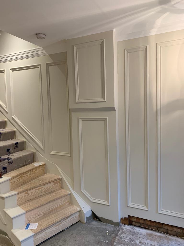 A staircase with bare wooden steps is partially covered in protective plastic and cardboard, hinting at an ongoing interior design project. The surrounding walls feature light cream paneling with decorative molding. A partially patched area is visible near the floor, exposing sections of the wall and cardboard.