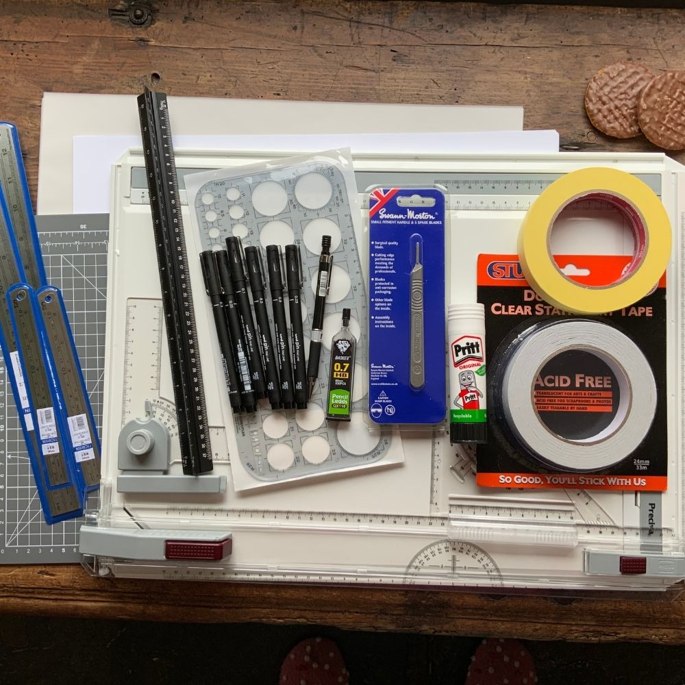 A drafting table with various architectural drawing tools, including a ruler, set squares, compass set, pens, mechanical pencils, eraser, utility knife, roll of clear tape and glue stick. Geometric stencils and a cutting mat are also visible on the table—perfect for students enrolled in an online interior design course.