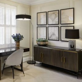 Get the look with a chic home office featuring a sleek black desk paired with a white chair, a floor lamp, and a fresh bouquet. A sideboard topped with a potted plant and decorative lamp sits against a wall adorned with six framed artworks. White shutters filter soft natural light.