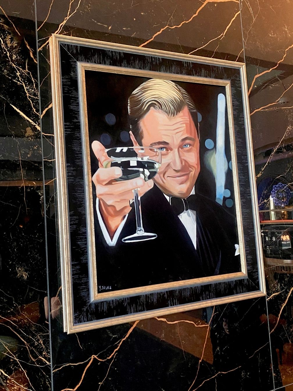 A framed painting of a man in a tuxedo holding up a cocktail glass in a toast graces the marbled wall of Diamond Suites, creating a sophisticated and elegant ambiance. The man has blonde hair and a slight smile as he looks directly at the viewer.