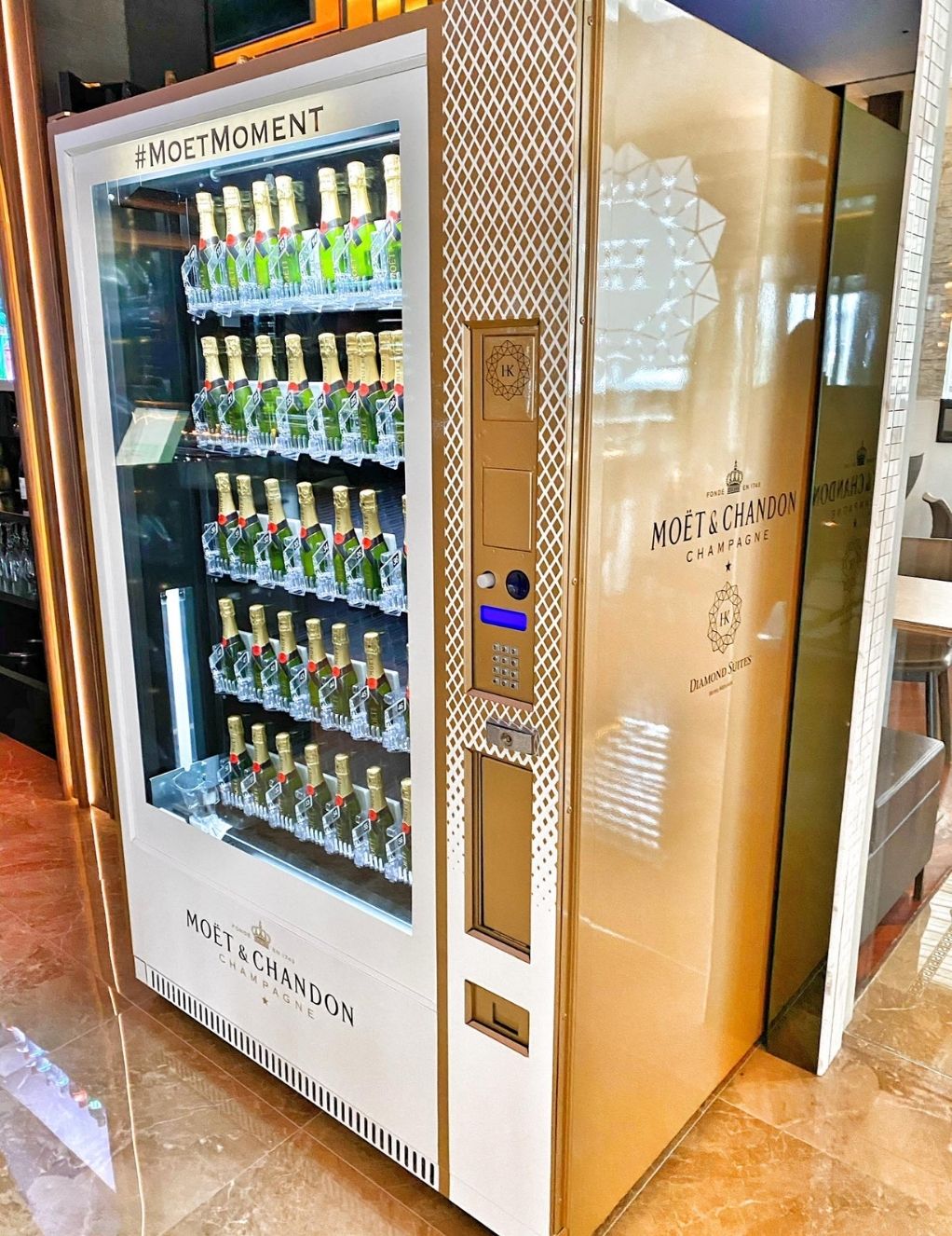 A sleek, gold-colored vending machine labeled "Moët & Chandon Champagne" dispenses mini bottles of Moët & Chandon. The machine features a digital interface and is branded with the hashtags "#MoetMoment" and "Diamond Suites." It rests on a shiny marble floor.