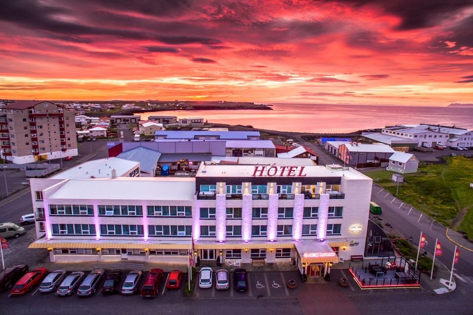 A brightly lit Diamond Suites hotel stands under a stunning sunset sky, with hues of pink, orange, and purple reflecting on the horizon. The hotel is situated in a coastal town, with buildings and the serene ocean visible in the background. Several cars are parked in front.