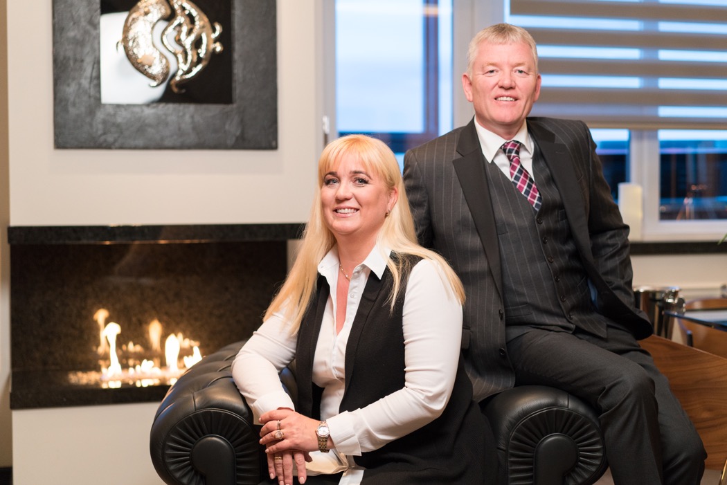 A man in a suit and a woman in business attire are posed indoors at Diamond Suites. The woman is seated on a black leather couch, and the man is sitting on its arm. Behind them is a modern fireplace with a small flame and artwork on the wall.