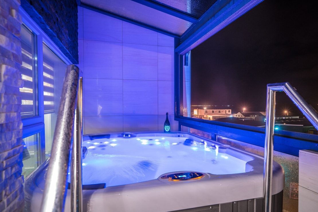 A view of an illuminated hot tub situated on a balcony at night in the luxurious Diamond Suites. The hot tub features blue lighting, bubbles, and occupies a tiled corner with a windowed wall on one side. City lights glow in the distance, adding sparkle to the background. A bottle sits on the tub’s edge.