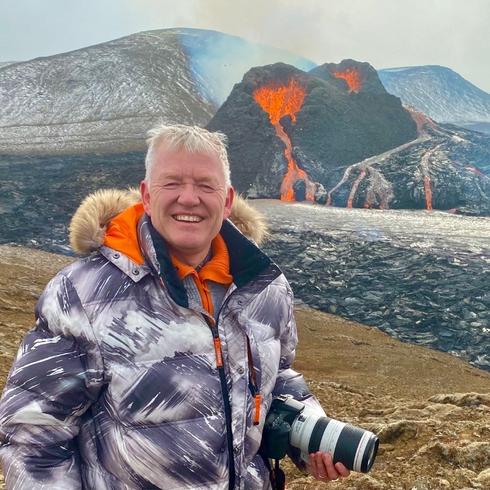 A person in a camouflage-patterned winter jacket stands smiling while holding a camera. Behind them, a volcanic eruption is visible with fiery lava flowing down the slope of a barren landscape. Snow-capped portions add contrast to the rugged terrain, creating an adventurous backdrop reminiscent of Diamond Suites' landscapes.