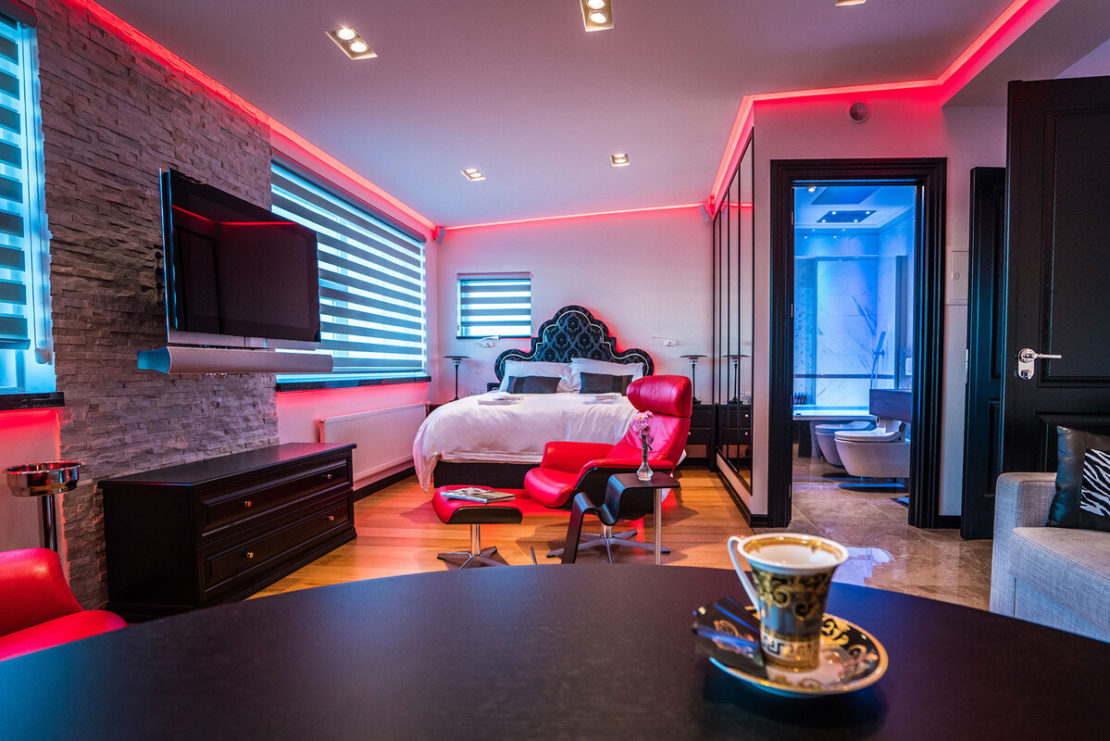 A modern bedroom at Diamond Suites boasts ambient pink lighting, a large bed, flat-screen TV, and a red lounge chair with matching ottoman. The room features a stone accent wall and a table in the foreground with a cup of coffee. There's an en-suite bathroom with a glass shower visible.