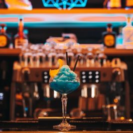 A blue slushy cocktail garnished with a twist of orange peel sits in a glass on the bar counter at Diamond Suites. The background features colorful, illuminated shelves stocked with various liquor bottles, while a coffee machine adds to the bar ambiance.