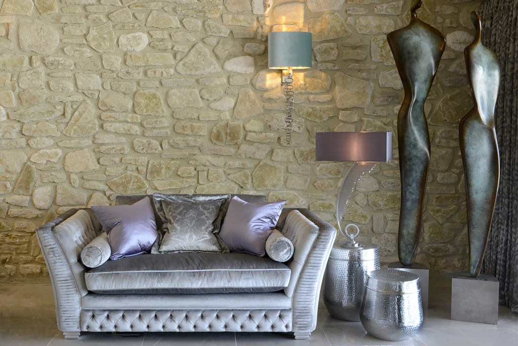 A light grey, tufted loveseat with metallic lilac cushions sits in front of a stone wall, adding personality to the décor. Two sculptural metal figures stand next to sculpted metallic side tables. Above the loveseat is a wall sconce with a light blue shade alongside a modern lamp on the side table.