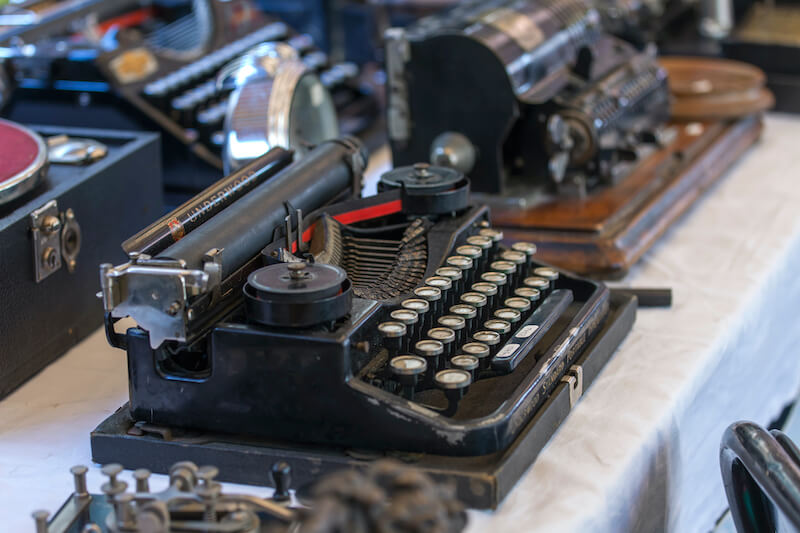 A vintage black typewriter displayed on a table, surrounded by various antique office equipment, adds personality to the setting. The typewriter has round keys and a red and black ribbon. The background shows more antique devices, including another typewriter and a machine with numbered dials.