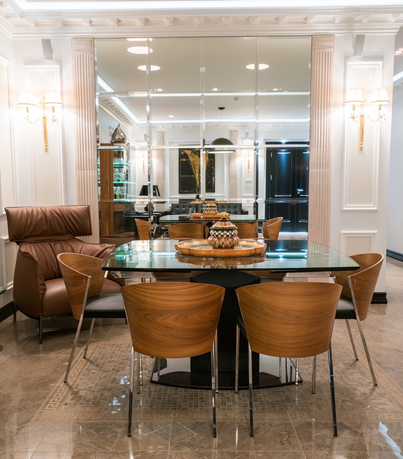 A modern dining room featuring a rectangular glass table surrounded by six wooden chairs, curated by Juliettes Trade. A brown leather armchair sits to the left. The room has mirrored walls with ornate white paneling, two wall sconces, and a tiled marble floor. A decorative centerpiece graces the table.