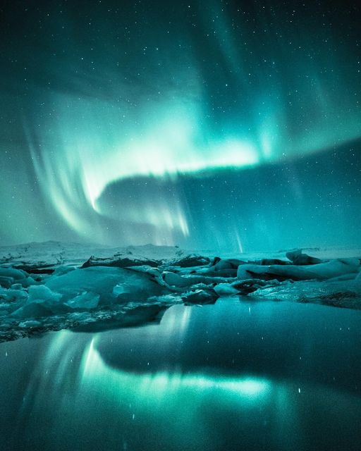 A vivid aurora borealis illuminates the night sky over an icy landscape with calm waters reflecting the green and blue lights. The stars are visible as tiny points of light in the background, creating a serene and mystical atmosphere, perfect for a luxury Iceland break.