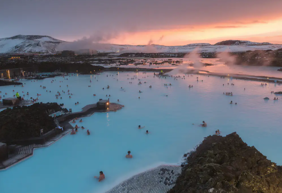 A panoramic view of the Blue Lagoon in Iceland at sunset, with numerous people enjoying a luxury Iceland break by swimming in the geothermal spa's milky blue waters. Snow-covered mountains and clouds of steam rise in the background, creating a serene and picturesque atmosphere.