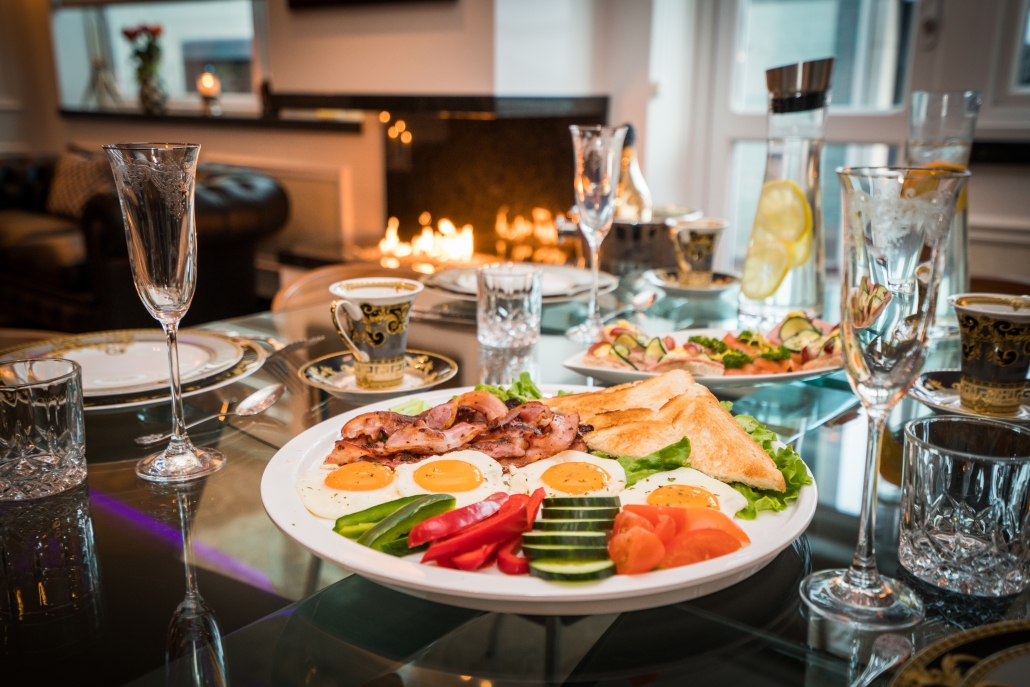 A lavish breakfast spread is laid out on a glass table at Diamond Suites. The main plate features fried eggs with crispy bacon, toast, and fresh vegetables like tomatoes and cucumbers. There are elegant cups and glasses, a water pitcher with lemon slices, and a fireplace in the background.