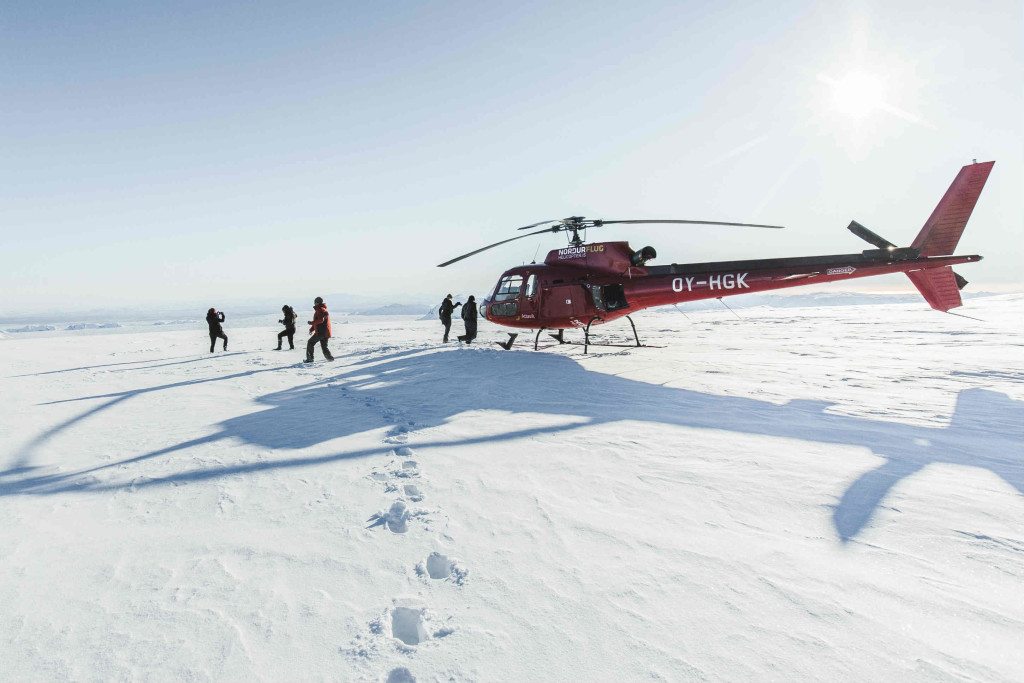 A red helicopter is parked on a vast, snowy landscape under a clear sky, part of a luxury Iceland break. Several people in dark winter clothing gather near the helicopter as the sun shines brightly, casting long shadows on the snow. Footprints lead away from the scene.