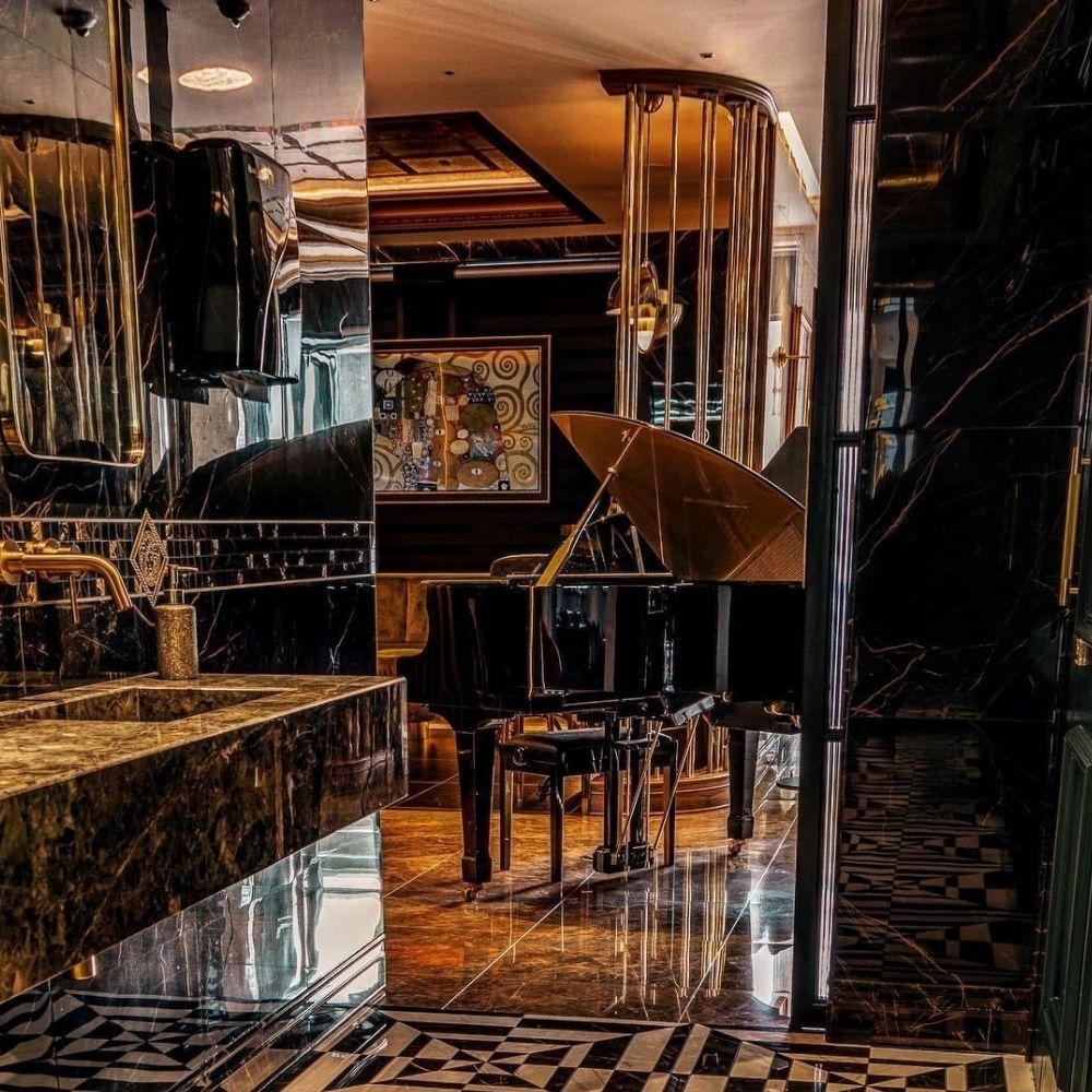 A luxurious indoor scene reminiscent of a luxury Iceland break, featuring a grand piano set near a black marble sink. The room is adorned with intricate black and gold decor, reflective surfaces, and a piece of modern art on the wall. The ambient lighting enhances the opulence of the setting.